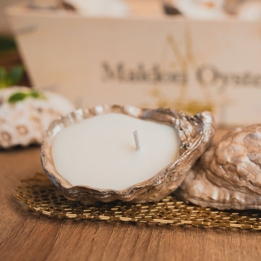 Candle inside an oyster shell on a wooden surface with 'Maiden Oyster' branding in the background.