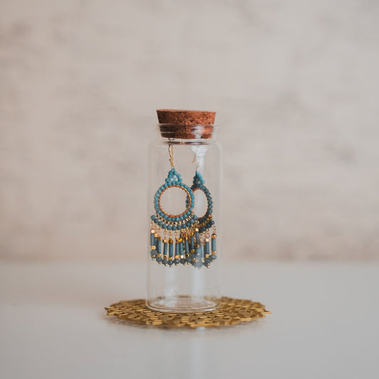 Decorative earrings in a glass bottle with cork lid on a white surface.